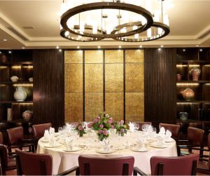An interior view of an opulent private dining room in Singapore at Li Bai Cantonese Restaurant. A large round table is set with 12 chairs under a sophisticated circular chandelier, flanked by built-in dark wood shelving and textured wall panels, reflecting a refined Cantonese fine dining atmosphere.